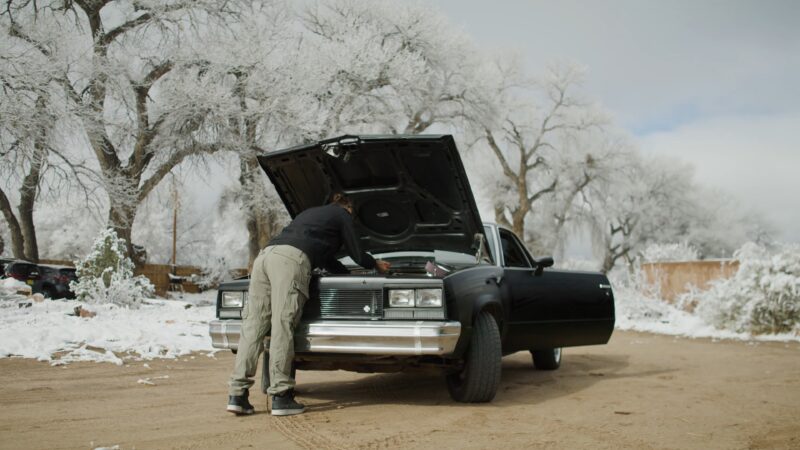 Rose B. Simpson leans into a black car with the hood open, inspecting the engine on a snowy day with bare trees in the background.
