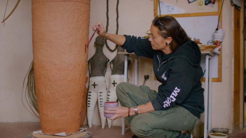 Rose B. Simpson kneels while painting a large, ceramic object indoors; decorated abstract figures are visible in the background.