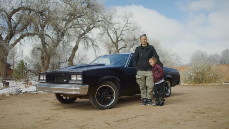 Rose B. Simpson and her child stand next to a black vintage car on a dirt road, with leafless trees and a cloudy sky in the background.