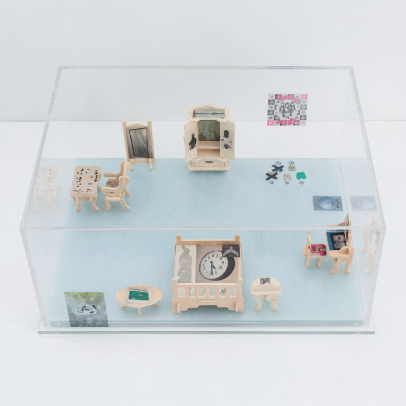 A clear box displays miniature wooden furniture with small objects and artwork on the walls and surfaces, arranged to resemble a living space.
