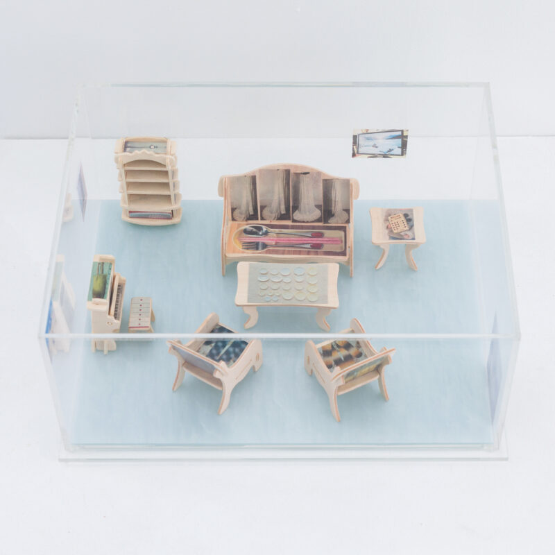 A clear box displays miniature wooden furniture with small objects and artwork on the walls and surfaces, arranged to resemble a living space.