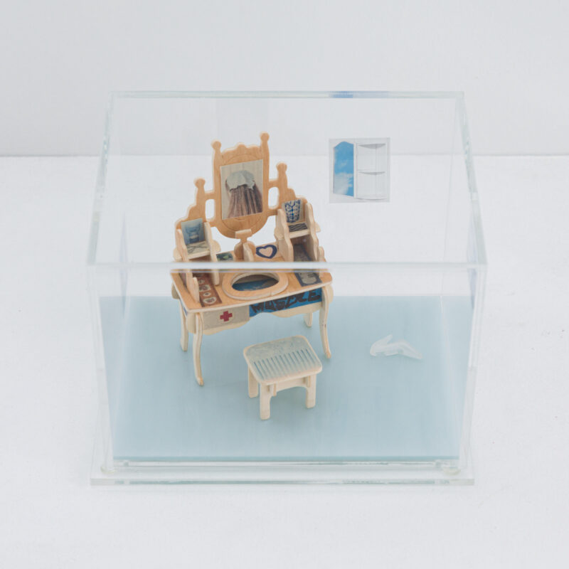 A glass box containing a miniature vanity table with a mirror, books, and small objects, with a tiny stool and a wall-mounted window.