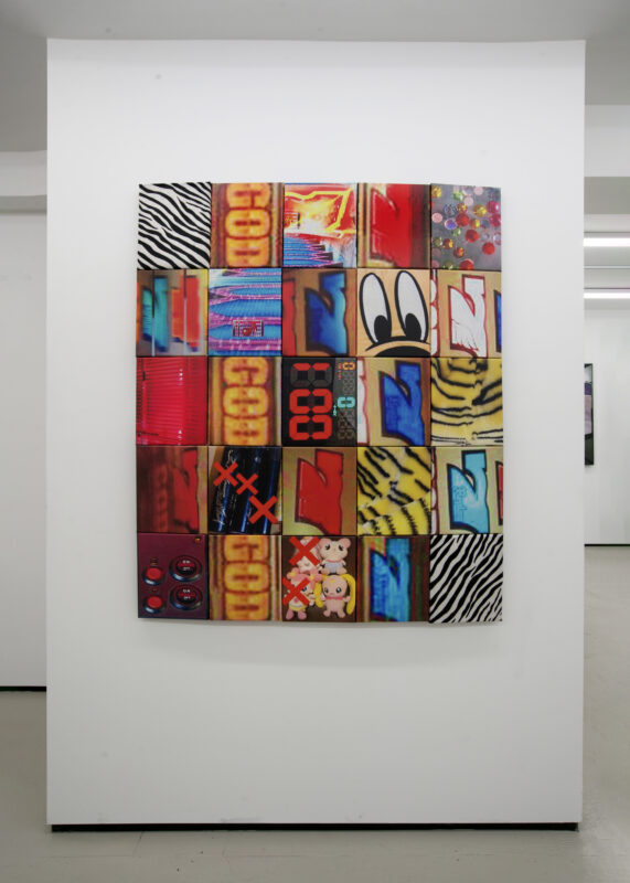A colorful, rectangular artwork composed of 24 blocks featuring cartoon eyes, numbers, patterns, and abstract designs hangs on a white gallery wall.