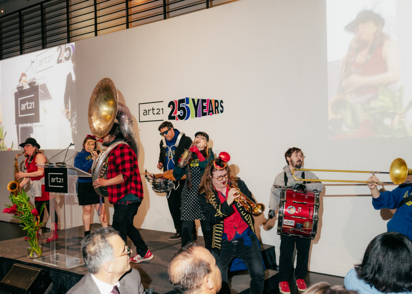 A brass band performs on stage at an Art21 25 Years anniversary event, with musicians playing various instruments and attendees watching in the foreground.