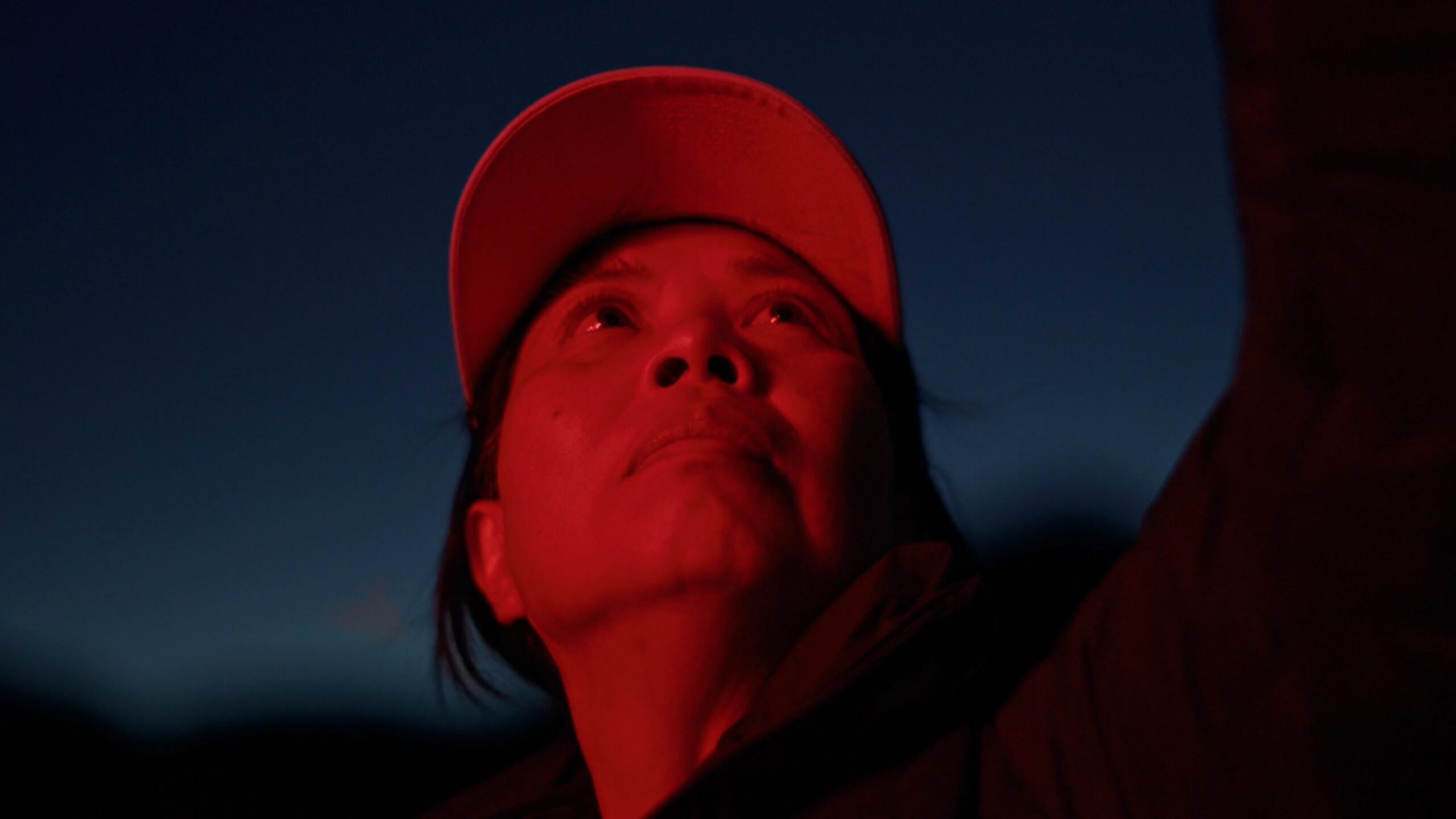 A person wearing a cap looks upward, illuminated by a red light against a dark sky.