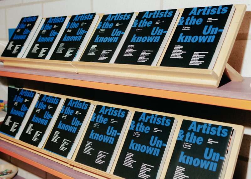 Several copies of a book titled “Artists & the Unknown” are displayed on two wooden shelves, arranged upright and side by side.