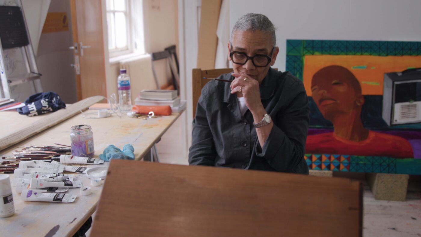 Lubaina Himid with short gray hair and glasses sits in an art studio, holding a paintbrush near her face, with art supplies and a colorful painting visible in the background.