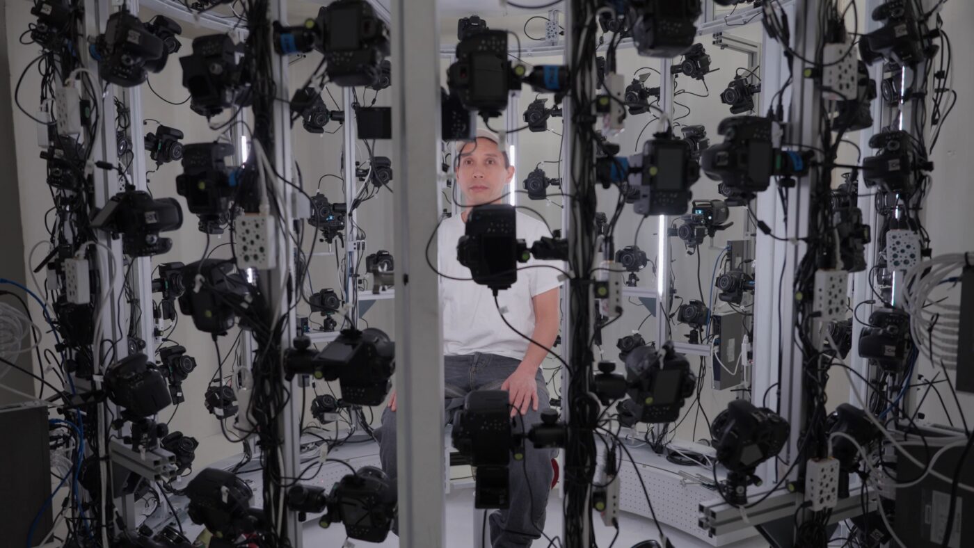 Josh Kline sits in the center of a room surrounded by many cameras mounted on metal frames, for a 3D scanning process.