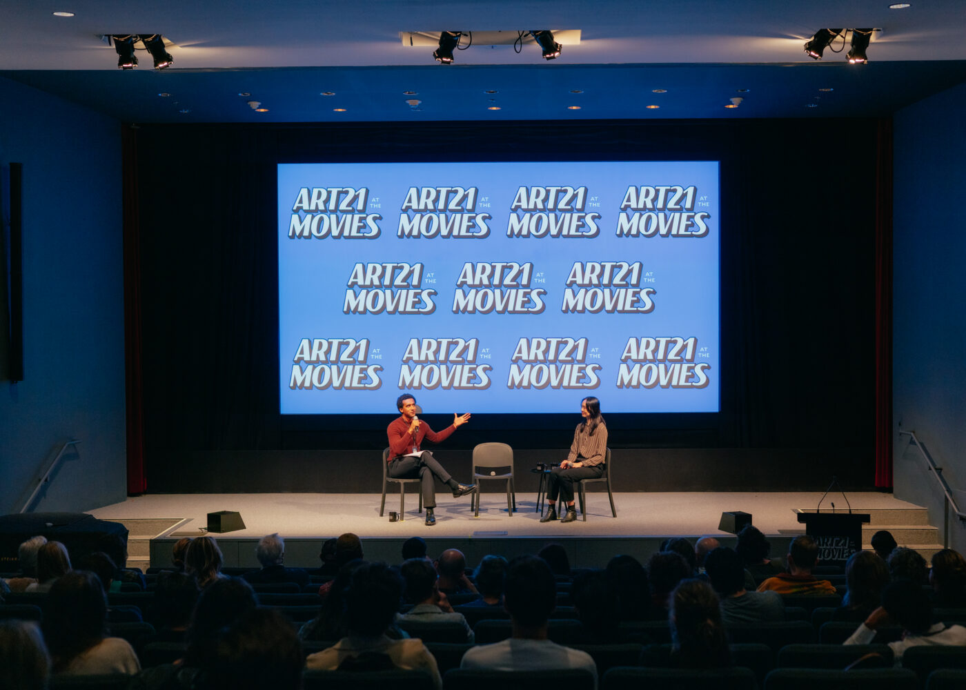Two people sit on stage having a discussion in front of an audience, with a large screen behind them displaying the text “ART21 at the MOVIES” repeatedly.
