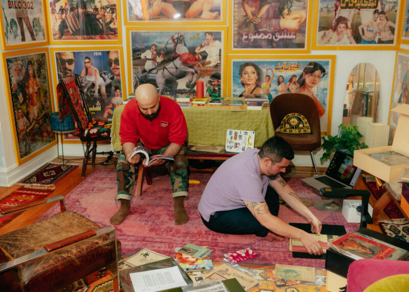 Two men organize and browse magazines and photos in a colorful room decorated with vintage posters and rugs.