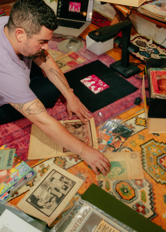 A person arranges vintage papers and photos on a patterned rug, surrounded by documents, a scanner, a laptop, and various books.