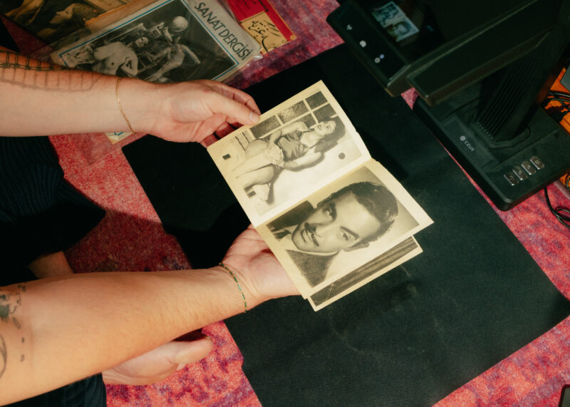 Hands holding vintage black-and-white photographs near a flatbed scanner, with magazines and other papers in the background on a pink carpet.