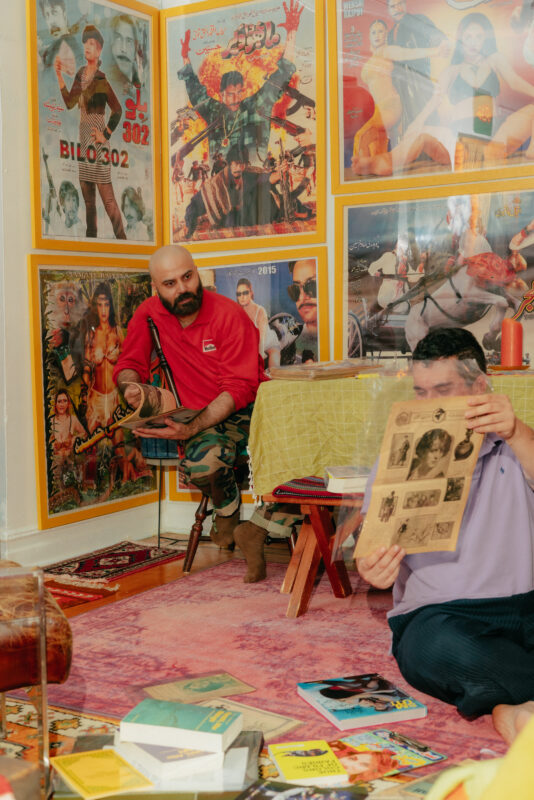 Two people sit in a colorful room with vintage movie posters on the walls; one person holds a booklet and the other looks on while sitting by a low table with scattered books and magazines.