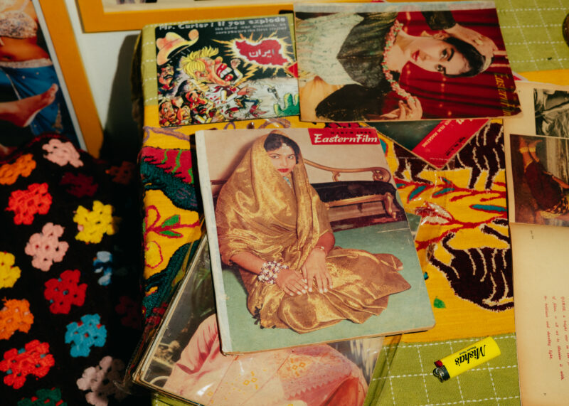 A stack of vintage magazines featuring women in traditional attire lies on a table with colorful crochet and a yellow lighter labeled “Misha's.”