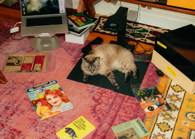 A cat lies on a black mat on a colorful rug, surrounded by books, magazines, a laptop, and other assorted items in a cluttered room.