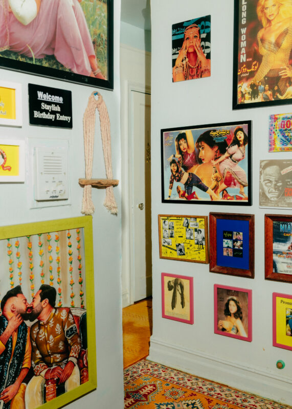 A white wall decorated with colorful movie posters and framed photos, with a woven wall hanging and a small hallway partially visible in the background.