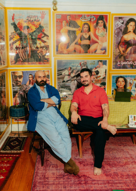 Two men sit side by side in a room decorated with colorful vintage movie posters covering the walls.