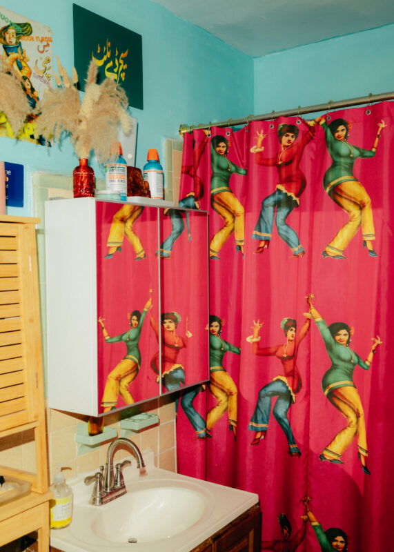 A bathroom with a pink shower curtain featuring dancing women in colorful outfits, a sink, a mirrored cabinet, and various decorations on light blue walls.