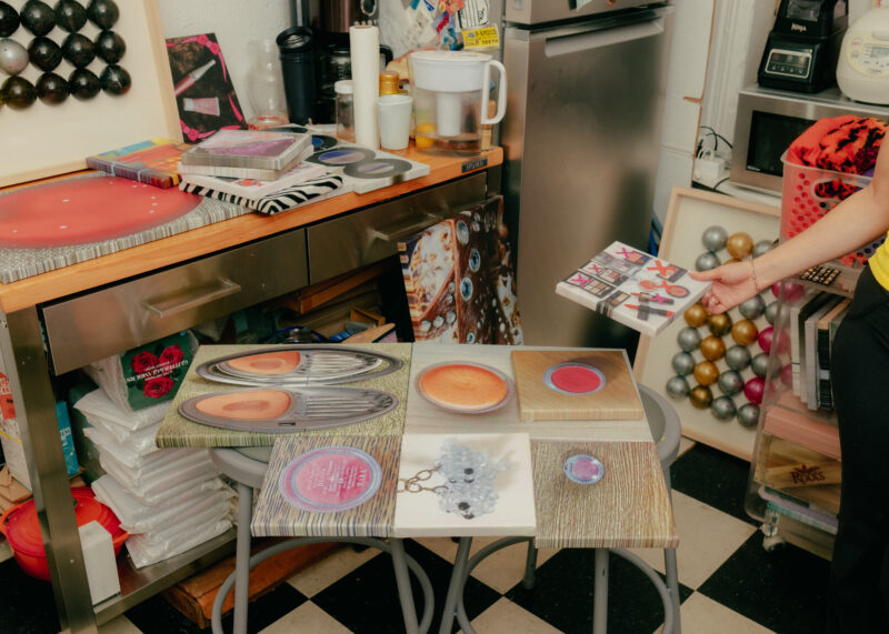 Person displaying small canvasses balanced on two stools; kitchen appliances and supplies are visible in the background.