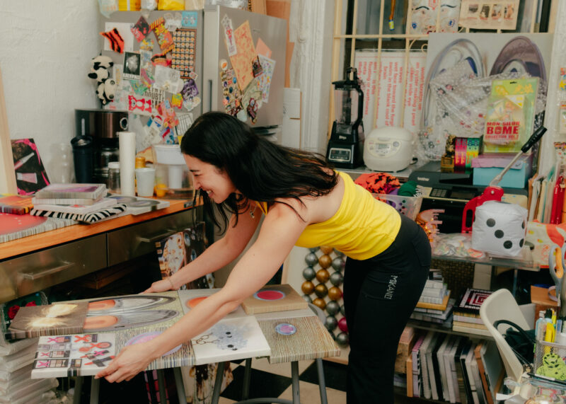 Sara Yukiko in a yellow tank top arranges colorful artwork on two tall stools in a cluttered, art-filled studio space.