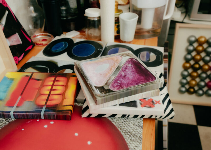 A colorful palette sits on top of books on a wooden surface, with various containers and objects in the background.