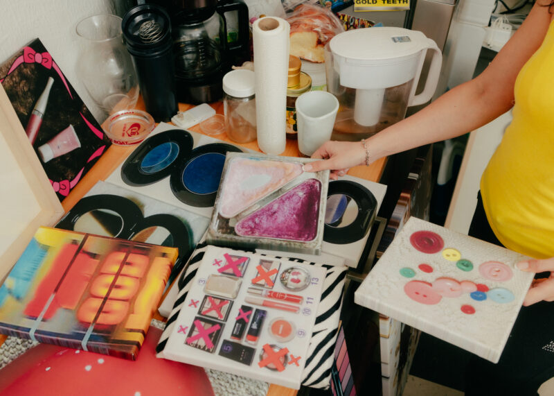 Person displaying colorful artwork and crafts on a cluttered kitchen counter with cups, appliances, and paper towels visible in the background.