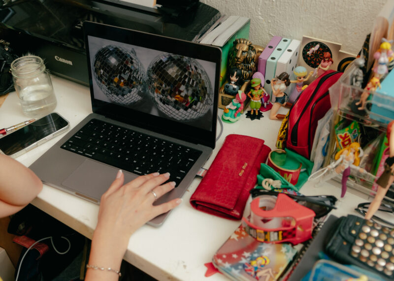 Person using a laptop at a cluttered desk with figurines, a red wallet, phone, and a glass of water. The laptop displays two side-by-side images of discos balls.
