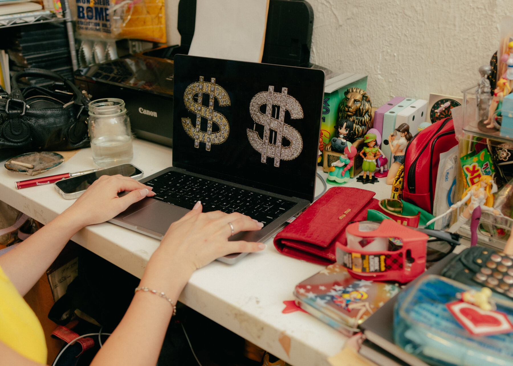 A person types on a laptop displaying two glittery dollar signs, surrounded by various personal items, figurines, and a water jar on a cluttered desk.