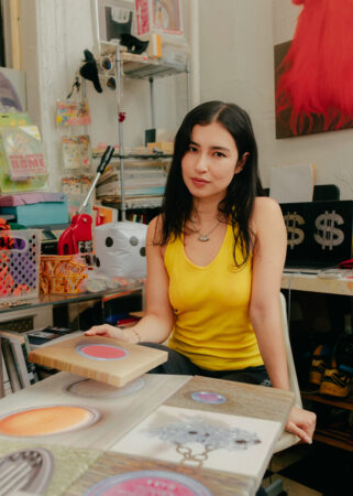 Sara Yukiko with long dark hair in a yellow tank top sits at a table with colorful circular artwork in a room filled with various art supplies.