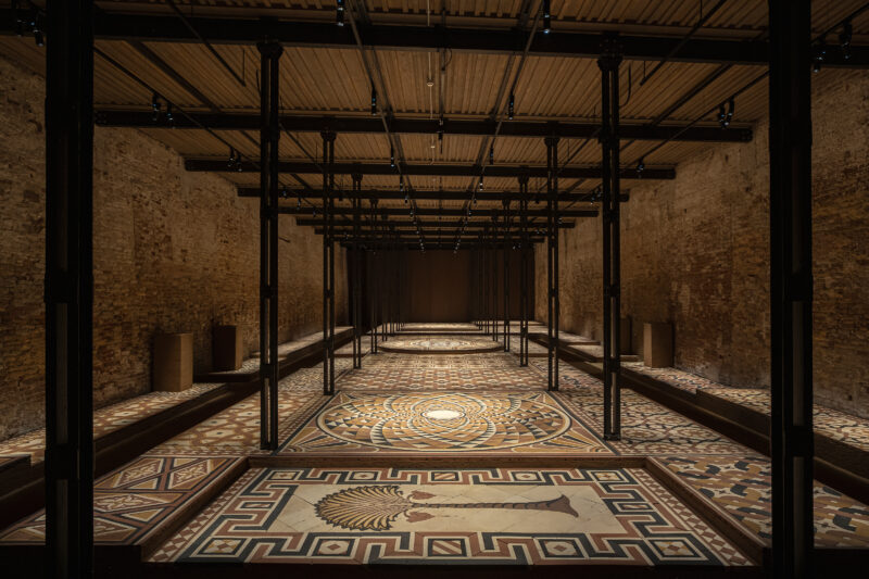 A long, narrow room featuring an intricate, multi-colored mosaic floor with geometric patterns and a central palm motif, enclosed by exposed brick walls and a grid of dark support columns.