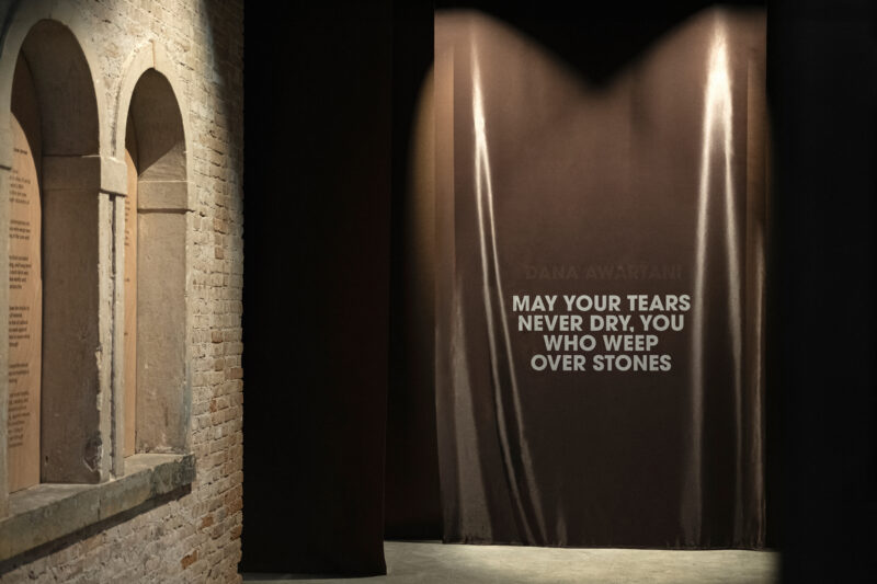 A dark fabric curtain displays the text “Dana Awartani: May Your Tears Never Dry, You Who Weep Over Stones,” next to an arched stone wall.