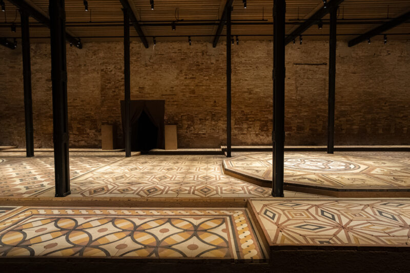 Large indoor space with exposed brick walls, support columns, and raised platforms featuring intricate, geometric tile patterns.