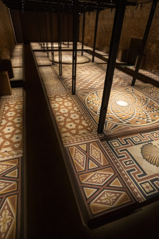 A large, high-angle view of an expansive floor covered in intricate, geometric mosaic tiles, with dark vertical support beams rising throughout the room.