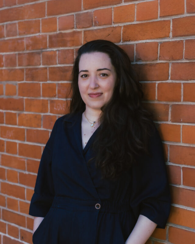 A woman with long dark hair posing against a red brick wall.