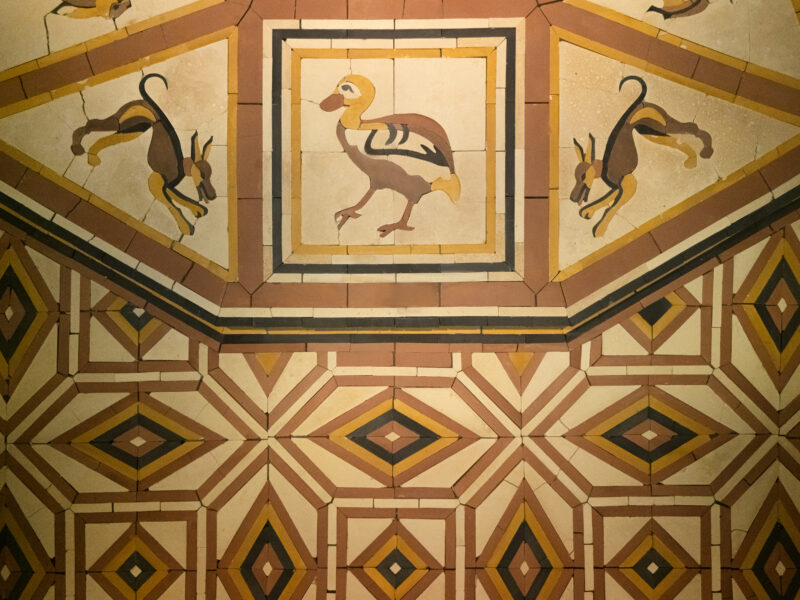 Floor tile mosaic featuring a central bird figure flanked by two stylized, leaping animals, set against a geometric pattern of diamonds and lines in tan, brown, and black.