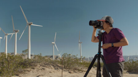 A person wearing a cap and purple shirt stands in a desert landscape, taking a photograph with a camera mounted on a tripod in front of several wind turbines.